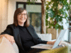 Alexandra Mert, Porträtfoto, eine junge Frau um die 30 Jahre auf einer Couch sitzend, mit Blazer und Laptop auf den Knien, schulterlange, braune Haare, offen, lächelnd, Brille tragend. Sie ist Expertin fur Behavioural Targeting