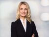 Porträt Frau mit langen blonden Haaren, leicht lächelnd, Ende 30, schwarzer Blazer, weiße Bluse, Business-Pose. Es handelt sich um Kathleen Rieser